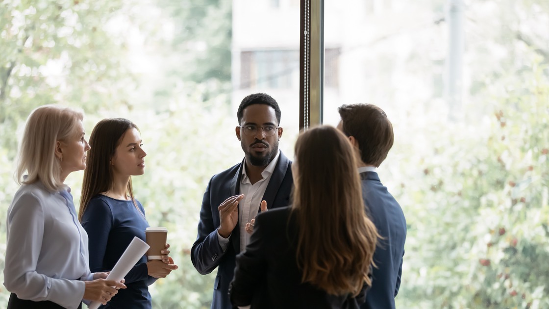 Focused multiracial businesspeople gather in office near window talk discuss business ideas in group together. Multiethnic diverse colleagues coworkers brainstorm at casual meeting. Teamwork concept.