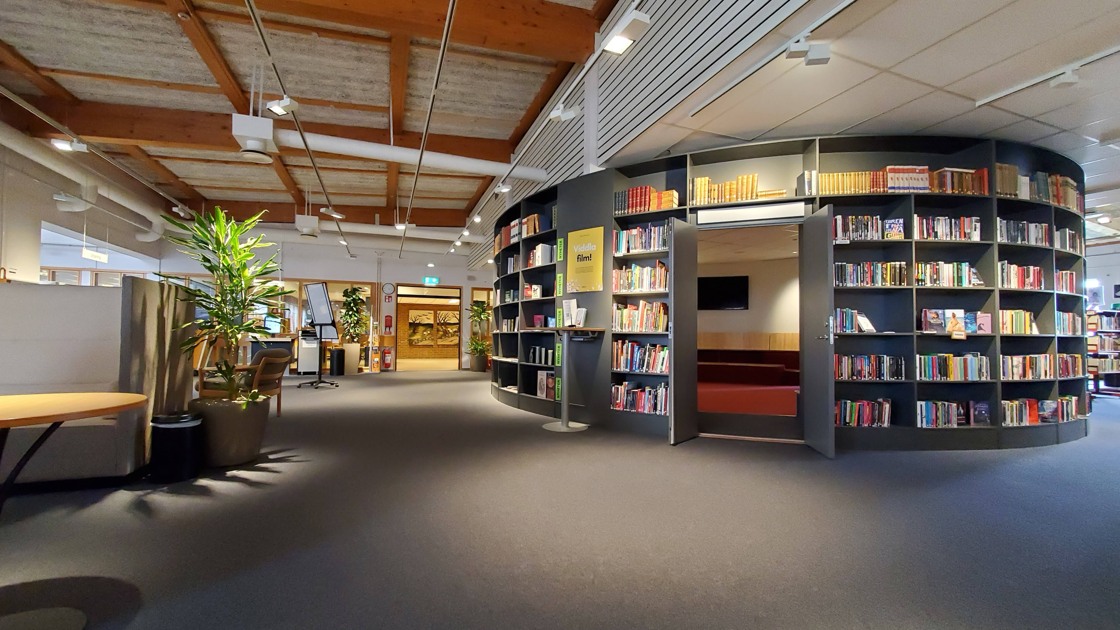 Miljöbild inifrån Tibro bibliotek. Stort öppet rum med mörk matta. Till höger i bild rotunda med bokhyllor och böcker.