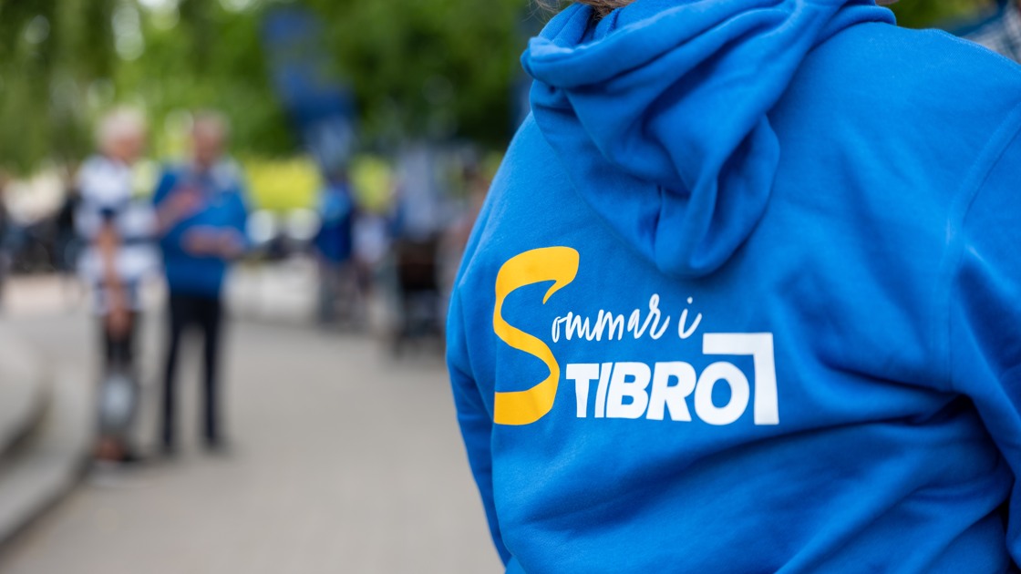På bilden syns ryggtavlan på en person som har en blå huvtröja på sig. På tröjan är ett tryck som säger "sommar i Tibro".