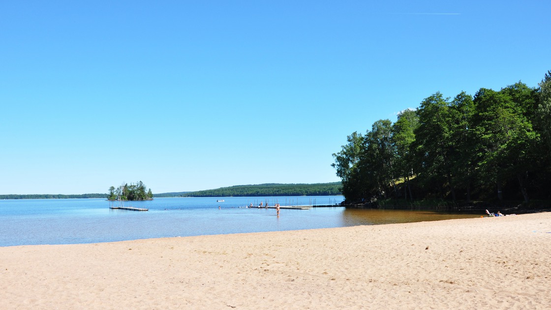 Solig dag vid Gamla Örlenbadet. I bild syns strand, vatten, Prästberget och Fläskön.