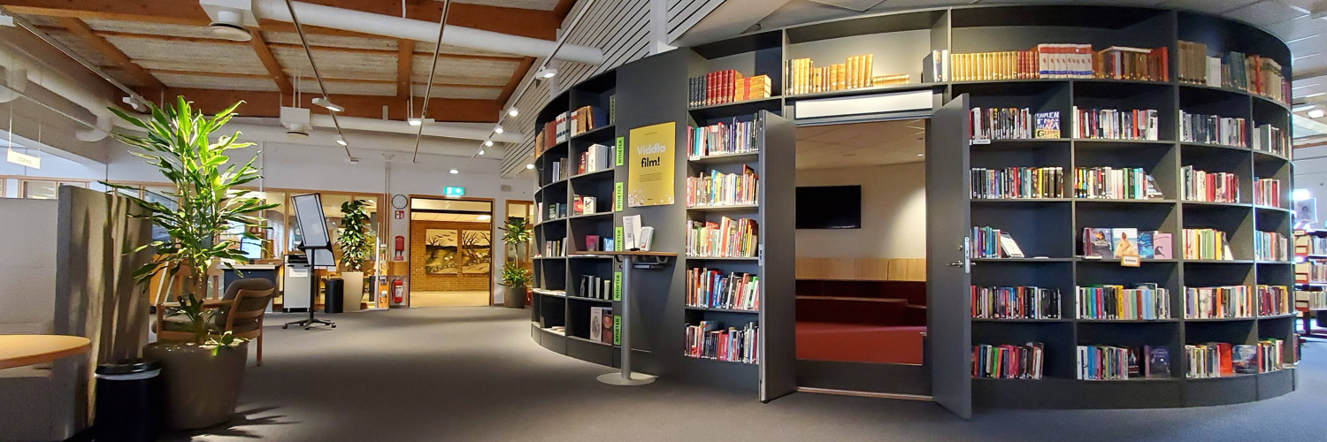 Miljöbild inifrån Tibro bibliotek. Stort öppet rum med mörk matta. Till höger i bild rotunda med bokhyllor och böcker.