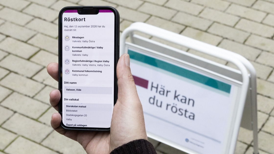 En person håller i en mobiltelefon med ett digitalt röstkort framför en skylt där det står Här kan du rösta.