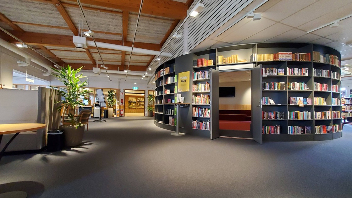 Miljöbild inifrån Tibro bibliotek. Stort öppet rum med mörk matta. Till höger i bild rotunda med bokhyllor och böcker.