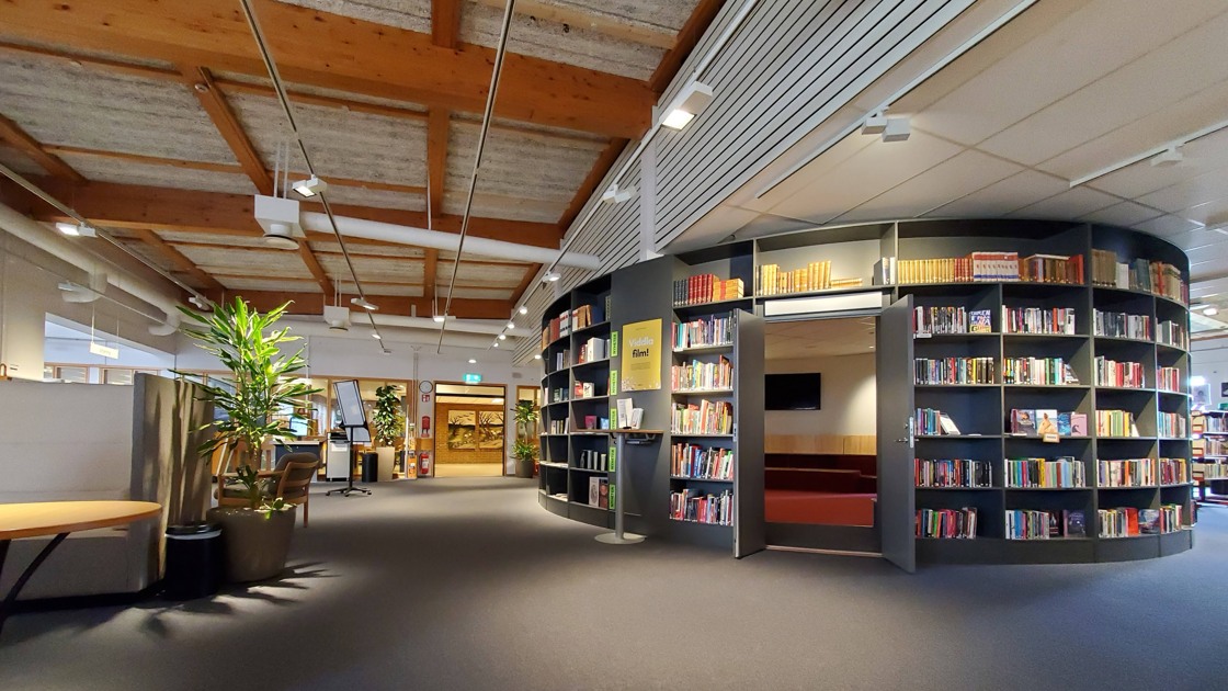 Miljöbild inifrån Tibro bibliotek. Stort öppet rum med mörk matta. Till höger i bild rotunda med bokhyllor och böcker.