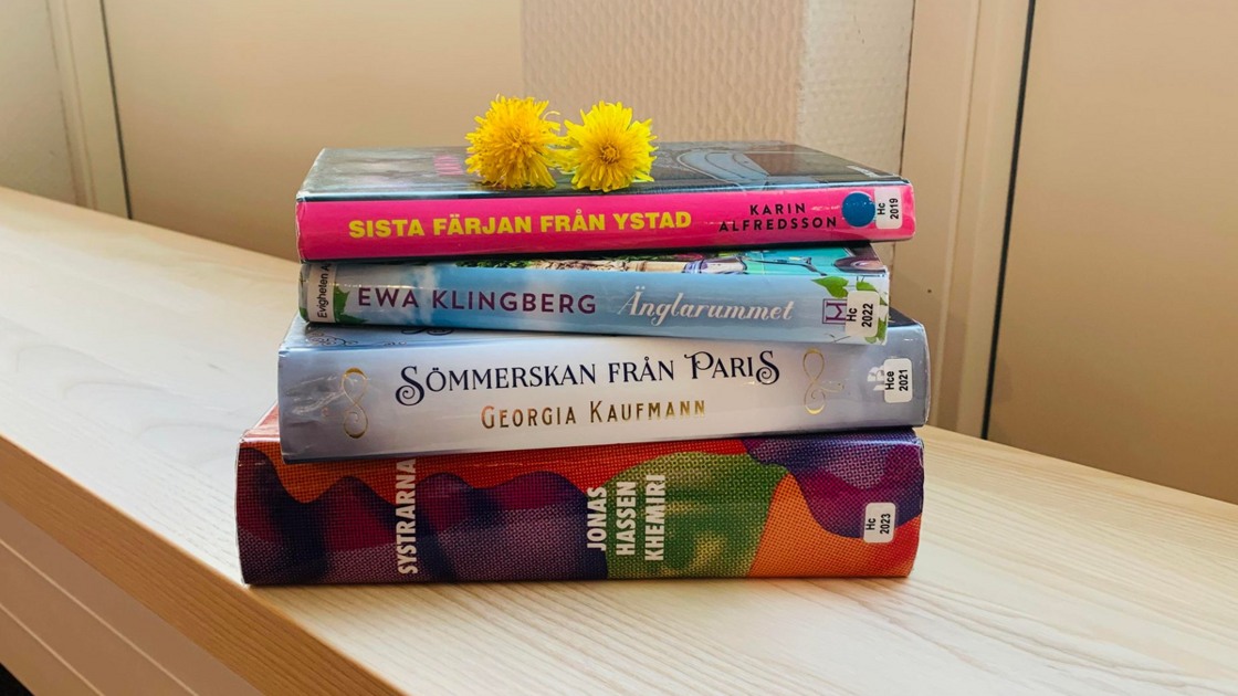 En bokhög med fyra böcker i olika färger och tjocklekar ligger på en ljus bänk. På boken högst upp ligger två maskrosor.