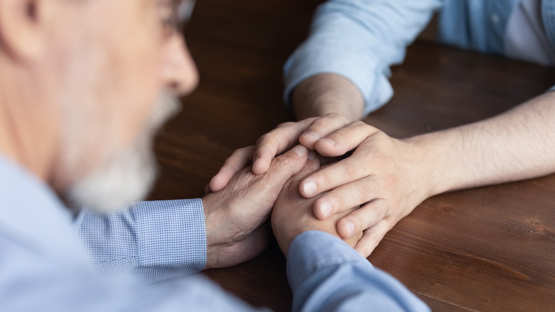 Supportive hands. Close up caring young man adult child caregiver sit at table facing old man retiree support comfort express empathy. Geriatric psychologist talk to elder patient on therapy session